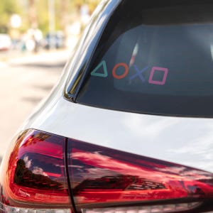 Puede incluir: La ventana trasera de un coche blanco presenta calcoman&iacute;as coloridas de s&iacute;mbolos de mando de videojuegos: un tri&aacute;ngulo verde, un c&iacute;rculo rojo, una X morada y un cuadrado rosa. La luz trasera del coche es visible debajo.