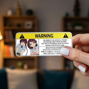 May include: A rectangular sticker with a yellow warning sign and the word "WARNING" in black. The sticker features anime-style illustrations of two girls and text warning against mocking waifus or anime, with potential removal from the vehicle.