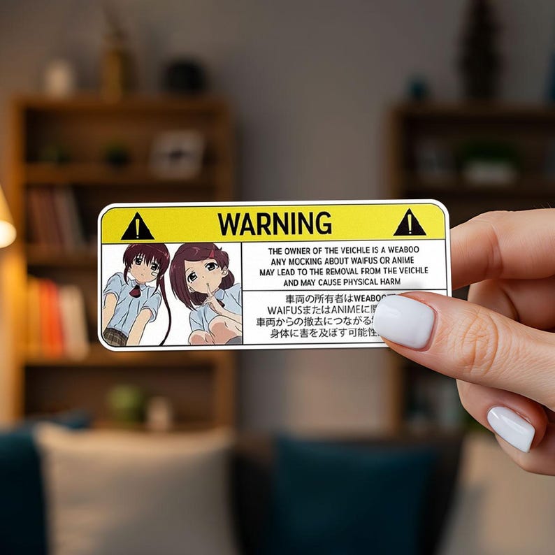 May include: A rectangular sticker with a yellow warning sign and the word "WARNING" in black. The sticker features anime-style illustrations of two girls and text warning against mocking waifus or anime, with potential removal from the vehicle.