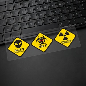 May include: Three yellow diamond-shaped stickers with black text and graphics. One reads "ALIEN ON BOARD" with an alien head, another says "AREA 51" with a biohazard symbol, and the last says "RADIOACTIVE MATERIALS" with a radiation symbol.