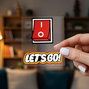 Puede incluir: Una pegatina transparente con un interruptor rojo, borde blanco y detalles blancos. El texto "LET'S GO!" está debajo en amarillo, rojo y blanco.