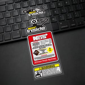 May include: Transparent stickers featuring black and white graphics and text, including "NOS Inside" and warning labels. The main label is red and white, detailing nitrous oxide mixture and safety warnings. The stickers are on a black keyboard.