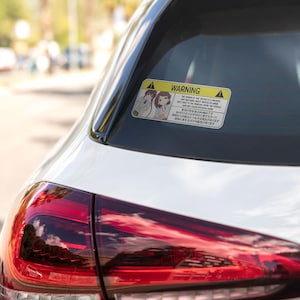 May include: A rectangular yellow and white warning sticker with anime-style illustrations and text, affixed to the rear window of a white car. The sticker features two cartoon characters and text in English and Japanese.