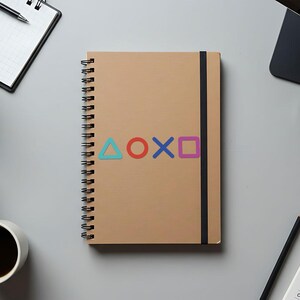 Puede incluir: Un cuaderno de espiral color beige con cierre el&aacute;stico negro. La portada muestra los s&iacute;mbolos de los botones del mando de PlayStation en azul, rojo, morado y verde. Se ven art&iacute;culos de oficina alrededor.