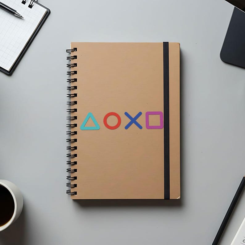 Puede incluir: Un cuaderno de espiral color beige con cierre el&aacute;stico negro. La portada muestra los s&iacute;mbolos de los botones del mando de PlayStation en azul, rojo, morado y verde. Se ven art&iacute;culos de oficina alrededor.