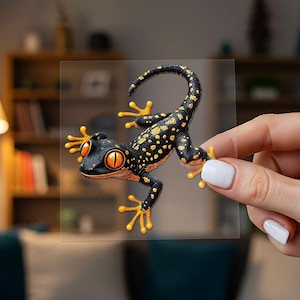 Peut inclure: Un autocollant transparent représentant un gecko noir avec des taches jaune vif et de grands yeux orange. Le gecko est représenté en position d'escalade avec la queue recourbée vers le haut, montrant une texture de peau détaillée.
