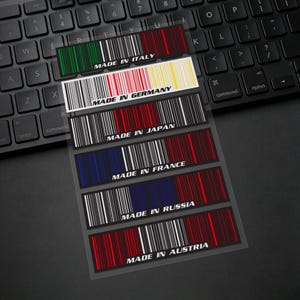 Puede incluir: Una hoja de pegatinas con diseños de códigos de barras y el texto "MADE IN" seguido de nombres de países. Las pegatinas utilizan los colores de las banderas de cada país, incluyendo Italia, Alemania, Japón, Francia, Rusia y Austria. Sobre un teclado.