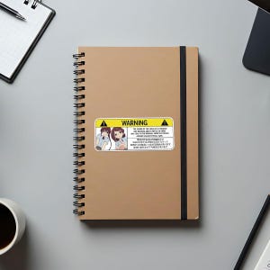 May include: A tan spiral-bound notebook with a black elastic band and a sticker that reads "WARNING" with an illustration of two anime-style girls. The sticker also contains text in English and Japanese.