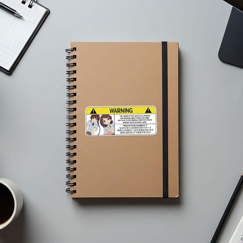 May include: A tan spiral-bound notebook with a black elastic band and a sticker that reads "WARNING" with an illustration of two anime-style girls. The sticker also contains text in English and Japanese.