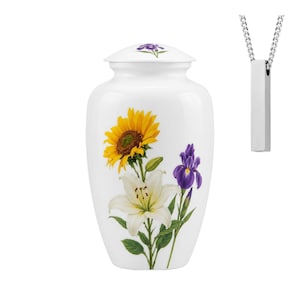 May include: A white cremation urn featuring a floral design of a yellow sunflower, a white lily, and a purple iris, with a matching floral motif on the lid. Beside the urn is a silver-toned rectangular pendant necklace on a matching chain.