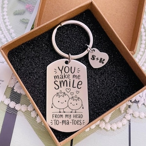 May include: A silver-toned metal dog tag keychain featuring the engraved text "YOU make me SMILE" above a graphic of two smiling tomatoes and the phrase "FROM MY HEAD TO-MA-TOES". A small heart-shaped charm with "S+K" is attached to the ring.