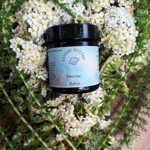 Pode incluir: Um pequeno frasco preto de bálsamo Yarrow Salve da Cosmic Butter com um rótulo azul-claro apresentando um ícone de planeta e texto. O frasco está aninhado entre cachos de pequenas flores brancas de mil-folhas com folhagem verde de samambaia.