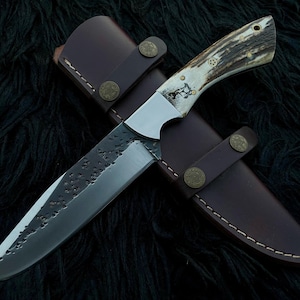 May include: A fixed-blade knife with a textured, dark-finished steel blade and a handle made of natural antler with brass pins. The knife is positioned over a brown leather sheath featuring white stitching and two circular brass-colored metal snaps.