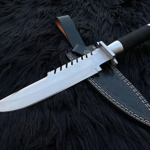 May include: A fixed-blade survival knife with a silver-toned steel blade featuring a serrated spine, a black cord-wrapped handle with a metallic pommel, and a black leather sheath with white contrast stitching and a belt loop attachment.