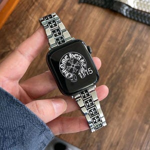 Puede incluir: Una correa de reloj plateada con una esfera negra. La esfera muestra el texto "Chrome Hearts" en blanco, junto con un diseño de cruz. La correa presenta un patrón de cruz repetido.