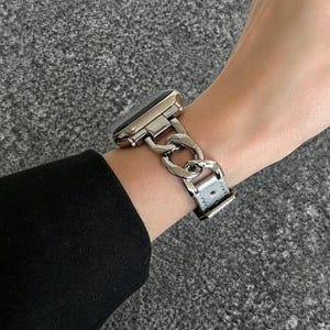 May include: A silver-toned watch with a chain-link band is worn on a wrist. The watch face is black. The band is a series of interlocking links. The watch band is attached to a gray leather strap.