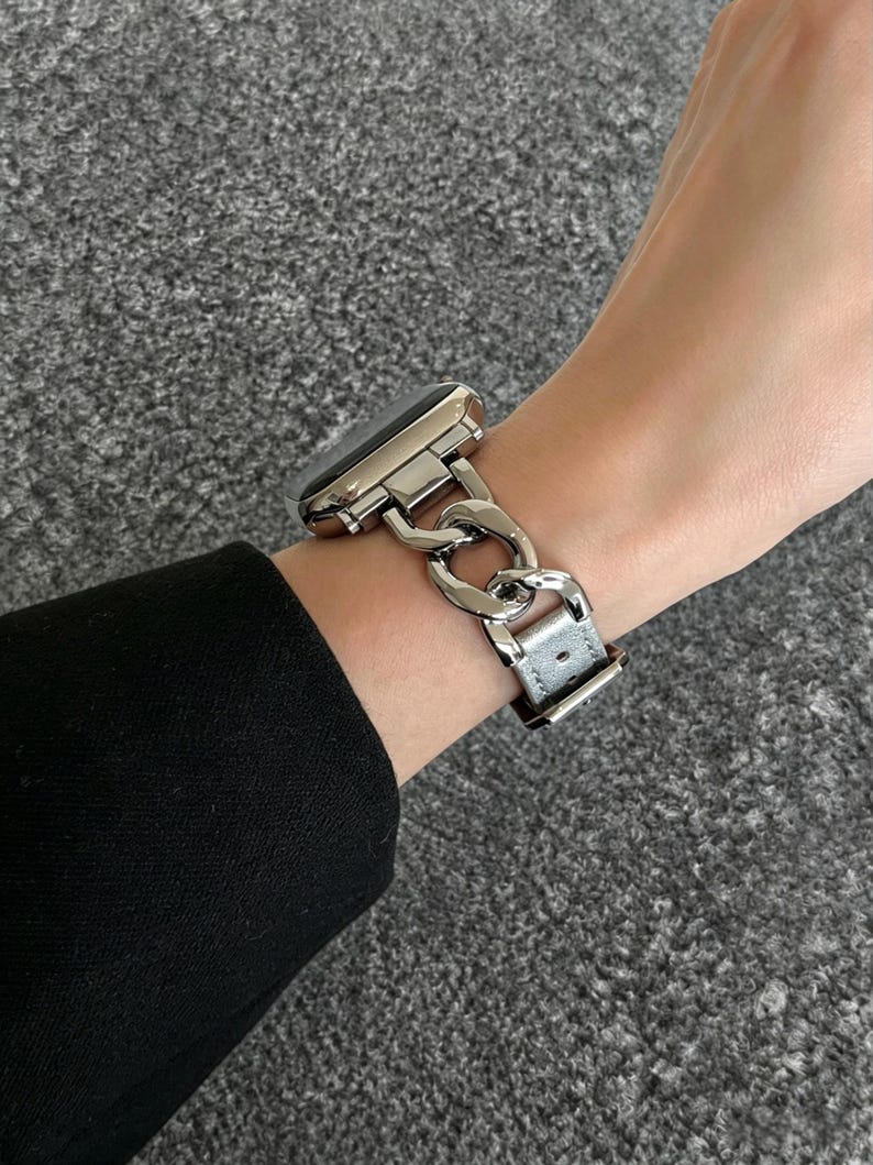 May include: A silver-toned watch with a chain-link band is worn on a wrist. The watch face is black. The band is a series of interlocking links. The watch band is attached to a gray leather strap.