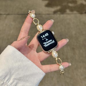 Könnte beinhalten: Eine goldfarbene Apple Watch mit einem Perlen- und Gold-Gliederarmband. Das Zifferblatt zeigt die Uhrzeit 14:48 und das Vivienne Westwood-Logo. Das Armband hat ovale und runde Goldglieder mit eingebetteten Perlen und kleinen Kristallen.