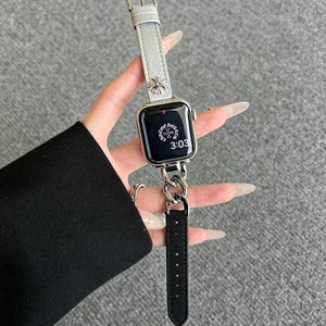 May include: A silver Apple Watch with a gray leather band and a silver chain attachment. The watch face displays the time 3:03. The band features a silver floral design.
