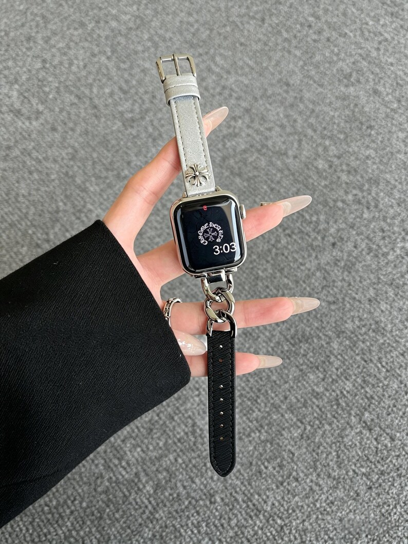 May include: A silver Apple Watch with a gray leather band and a silver chain attachment. The watch face displays the time 3:03. The band features a silver floral design.