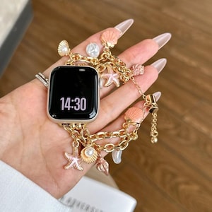 May include: A gold-toned smartwatch band featuring a chain-link design adorned with multiple charms, including pink seashell shapes, white pearls, and starfish figures. The watch face is black and displays the time 14:30 in white digital numerals.