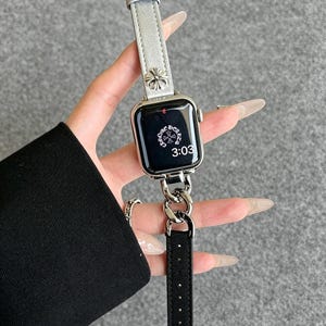 May include: Silver Apple Watch with a silver leather band and a black leather strap. The watch face displays the time as 3:03. The band features a decorative floral design.