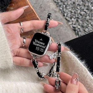 Puede incluir: Reloj inteligente plateado con pantalla negra que muestra "Vivienne Westwood" y la hora 15:15. El reloj tiene una correa de cuero negro con eslabones de cadena plateados y dijes de mariposa. El reloj está en las manos de una persona.