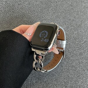 May include: A silver Apple Watch with a chain link band and a silver leather strap. The watch face displays the time as 2:49. The watch is held against a gray background.