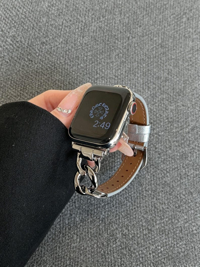 May include: A silver Apple Watch with a chain link band and a silver leather strap. The watch face displays the time as 2:49. The watch is held against a gray background.