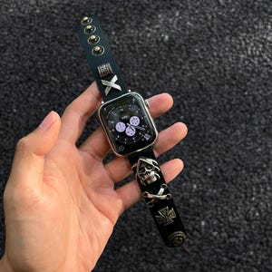Puede incluir: Correa de cuero negro para Apple Watch con adornos plateados de calaveras y huesos cruzados. La correa también presenta remaches plateados y una caja de reloj plateada. La esfera del reloj muestra la hora y otros datos.