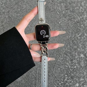 May include: Silver Apple Watch with a silver leather band and a silver chain link band. The watch face displays the time as 2:48. The band features a decorative cross design.
