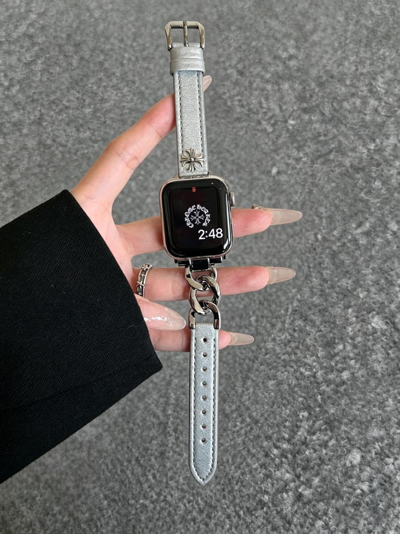May include: Silver Apple Watch with a silver leather band and a silver chain link band. The watch face displays the time as 2:48. The band features a decorative cross design.