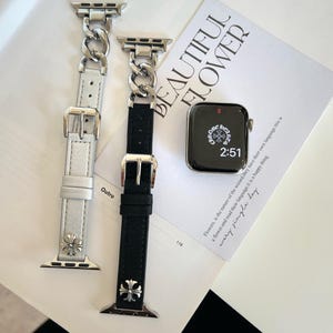 May include: Two watch bands with silver-tone chain links and leather straps, one white and one black, are displayed. Each band has a silver buckle and a decorative cross. A smartwatch with a black screen and the time 2:51 is also visible.