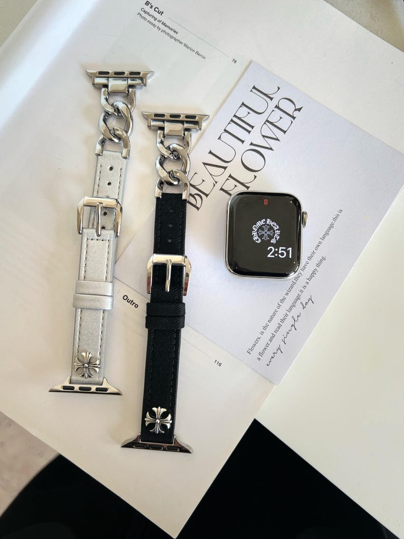 May include: Two watch bands with silver-tone chain links and leather straps, one white and one black, are displayed. Each band has a silver buckle and a decorative cross. A smartwatch with a black screen and the time 2:51 is also visible.