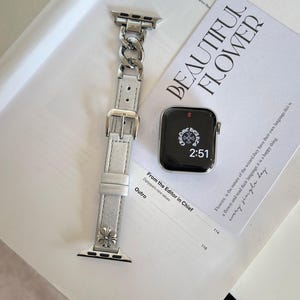 May include: A silver Apple Watch with a black screen displaying the time 2:51, next to a silver and white watch band. The band features a chain link design and a buckle. A magazine with the words "BEAUTIFUL FLOWER" is in the background.