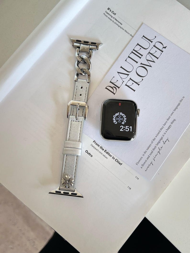 May include: A silver Apple Watch with a black screen displaying the time 2:51, next to a silver and white watch band. The band features a chain link design and a buckle. A magazine with the words "BEAUTIFUL FLOWER" is in the background.
