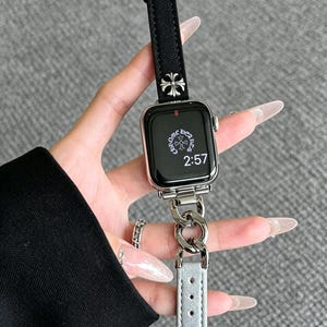 May include: Silver Apple Watch with a black fabric band and a silver chain link band. The watch face displays the time as 2:57. The band features a silver cross design.