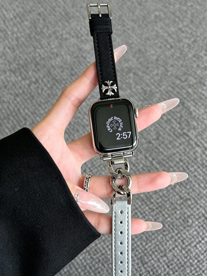 May include: Silver Apple Watch with a black fabric band and a silver chain link band. The watch face displays the time as 2:57. The band features a silver cross design.