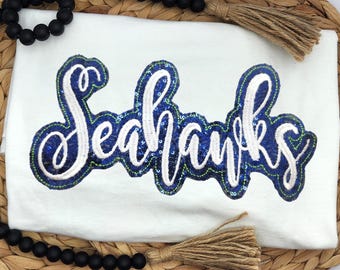 May include: White t-shirt featuring the word "Seahawks" in a blue and white sequined design. The shirt is presented on a woven placemat, with black beads and tassels.