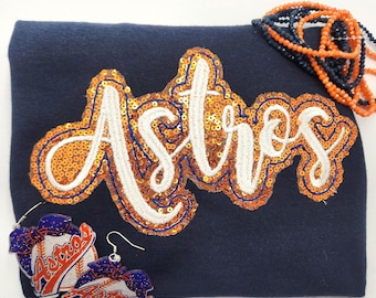 Puede incluir: Una sudadera azul marino con la palabra Astros en letra blanca y borde de lentejuelas naranjas. Un par de pendientes de béisbol con lazos azules y un collar de cuentas de varias hebras en naranja y azul marino se muestran encima.