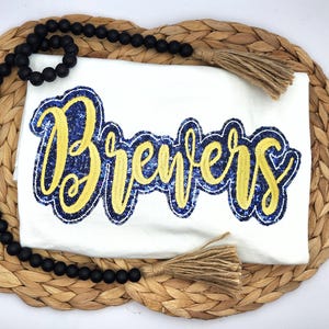 May include: A white t-shirt with the word "Brewers" in yellow script with a blue outline. The shirt is displayed on a woven placemat with a black beaded garland and jute tassels.