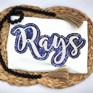 May include: A white t-shirt featuring the word Rays in a cursive font. The lettering is constructed with blue sequin fill and a thick white embroidered border. A strand of black wooden beads with tan jute tassels is draped over the shirt.