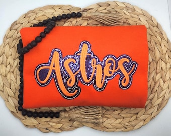 May include: A bright orange sweatshirt featuring the word Astros embroidered in a cursive font. The lettering is outlined in a dark purple sequined border with a lighter orange thread fill. A string of black wooden beads with tan tassels is draped.