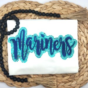 May include: A white t-shirt featuring the word Mariners in dark blue script embroidery, surrounded by a bright turquoise sequin border. The shirt is accented by a strand of matte black wooden beads with tan jute tassels at each end.