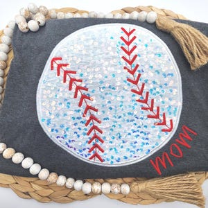 May include: A dark gray t-shirt featuring a large circular baseball graphic filled with iridescent white and blue sequins. The baseball design includes red stitched embroidery details and the word mom embroidered in red script on the lower right.