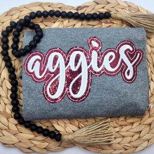 May include: A heather gray t-shirt featuring the word aggies in white script lettering with a dark red sequined outline. The shirt is draped over a round woven natural fiber mat and is accented by a strand of matte black wooden beads.