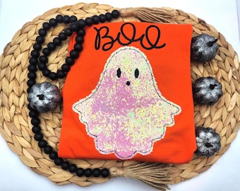 May include: An orange t-shirt featuring a large ghost applique covered in iridescent white and pink sequins. The word BOO is printed in black script above the ghost. The shirt is surrounded by black wooden beads and three small silver pumpkins.