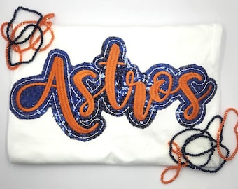 May include: A white t-shirt featuring the word Astros in orange script lettering, outlined with dark blue sequins. The design is surrounded by two loose strands of orange and dark blue beads, one positioned at the top left and one at the bottom right.
