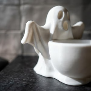 May include: A white 3D printed ghost figurine featuring a hollowed-out bowl section in the front. The ghost has an elongated head with two oval eye sockets and an open mouth, with arms extended outward to the sides, forming a small container.