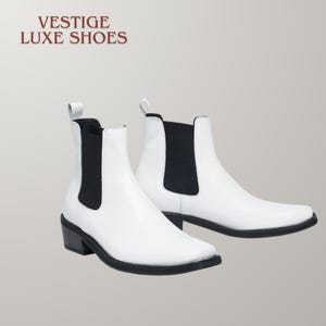 May include: A pair of white Chelsea boots with black elastic side panels and black soles. The text VESTIGE LUXE SHOES is printed in the upper left corner. These ankle boots feature a pull tab at the back and a low block heel for a clean look.